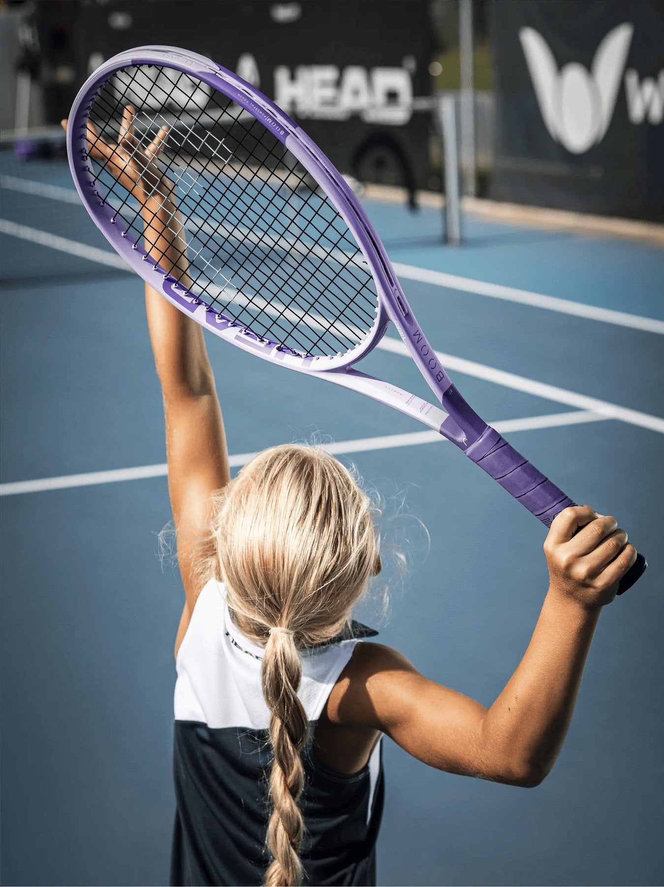 A young child with blonde hair holds up a HEAD Boom Jr 26 Alternate 2026 Junior Tennis Racket on a blue court, ready to serve or celebrate under the sun.