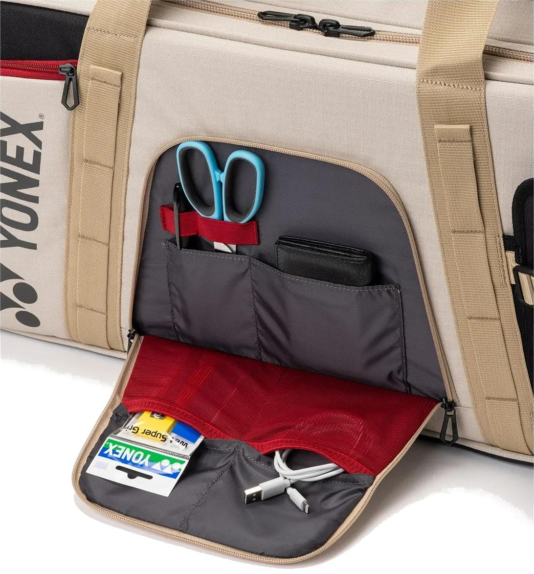 The Yonex BA72631WEX Gearlogic Tournament Tennis Bag in beige features an open front pocket with a red and gray lining, neatly storing blue scissors, pens, a black wallet, cards, USB cable, and mesh compartments—perfect for tournaments.