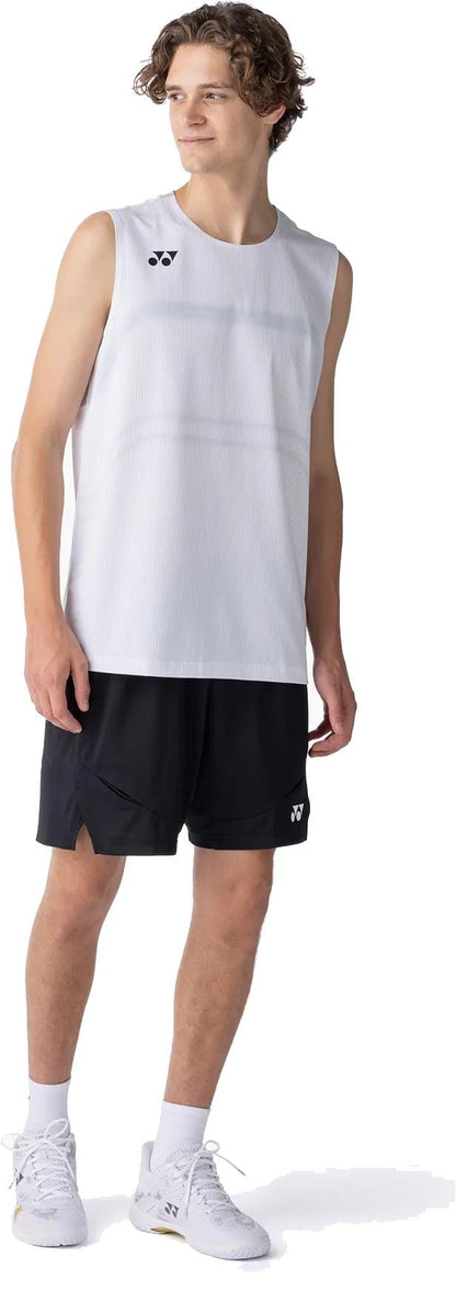 A young man with wavy hair wears a sleeveless white athletic shirt, Yonex 15239YX Knit Mens Tennis Shorts in black, and white sneakers while standing and looking to the side against a plain white background.