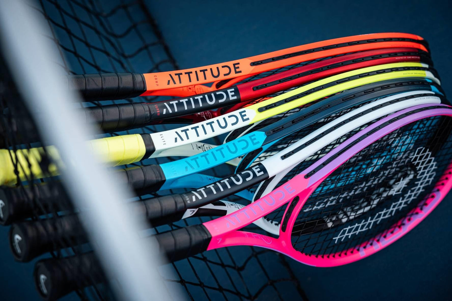 HEAD MX Attitude COMP Tennis Rackets by HEAD, featuring "ATTITUDE" on the throat, are stacked together at a tennis net on a blue court. These lightweight rackets come in bright red, orange, yellow, gray, blue, and pink shades.
