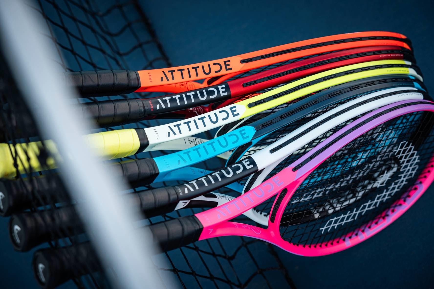 HEAD MX Attitude COMP Tennis Rackets by HEAD, featuring "ATTITUDE" on the throat, are stacked together at a tennis net on a blue court. These lightweight rackets come in bright red, orange, yellow, gray, blue, and pink shades.