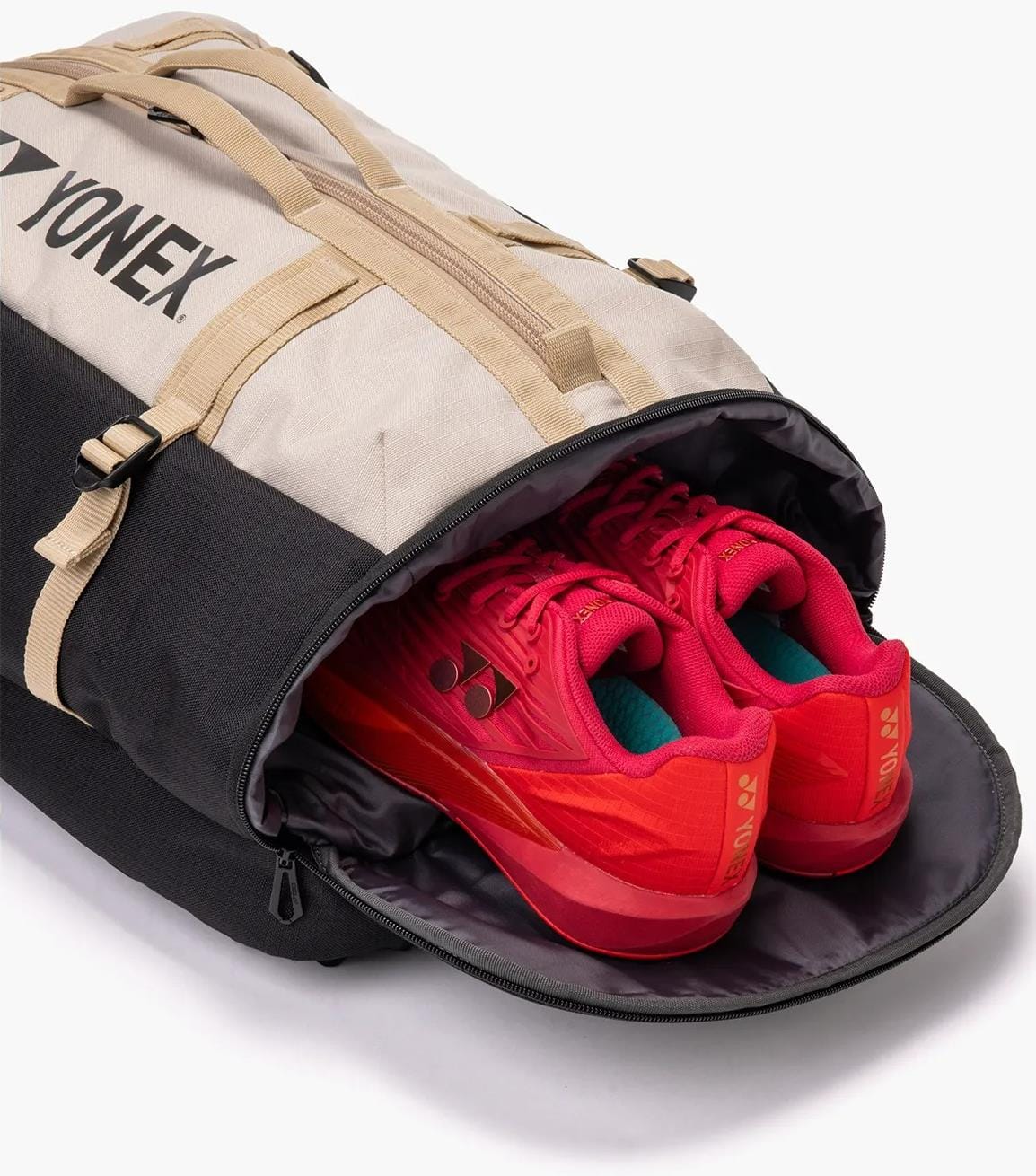 A Yonex BA72612EX Gearlogic Tennis Backpack in beige with a partially open zippered compartment reveals a pair of bright red athletic shoes inside.
