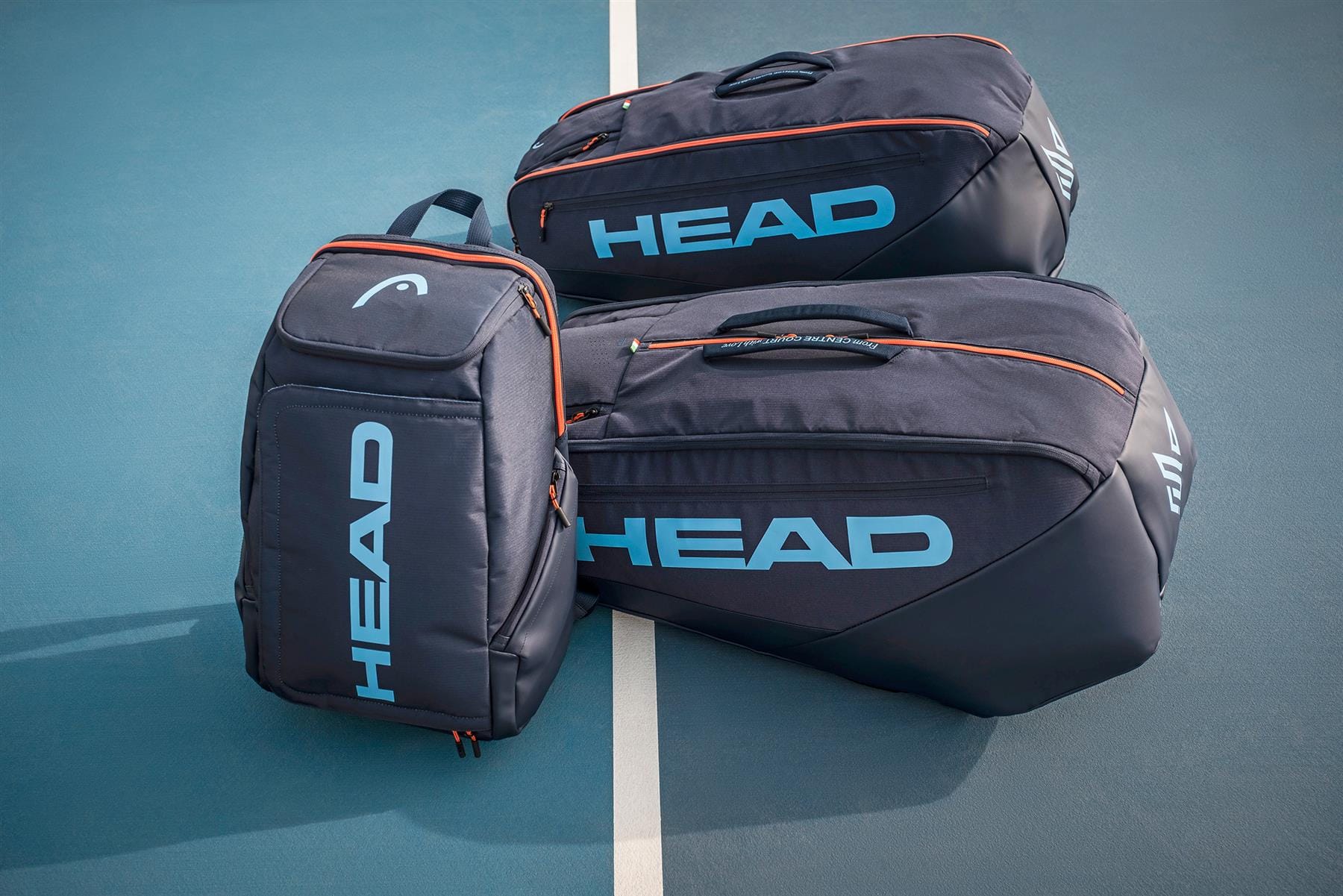 Three HEAD Pro 9 Racket Tennis Bag L - Navy bags, featuring light blue logos and orange accents, are displayed on a blue court near a white line—ideal for those seeking a stylish, climate-controlled tennis bag like Jannik Sinner’s.