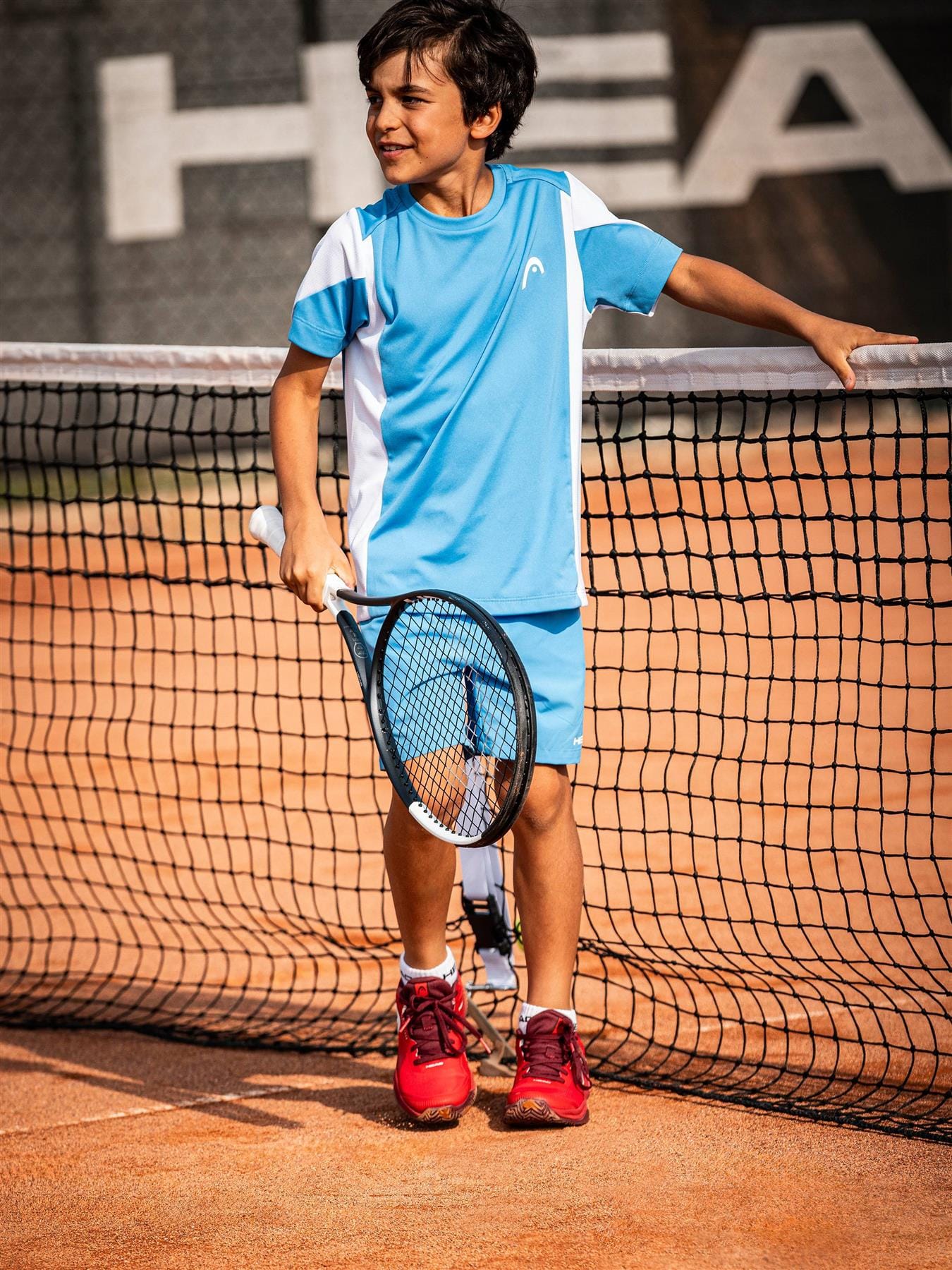A young boy in a blue sports outfit smiles and leans against the net on a clay court, holding his HEAD Speed Junior 26 2026 Tennis Racket. The background displays part of a tennis banner.