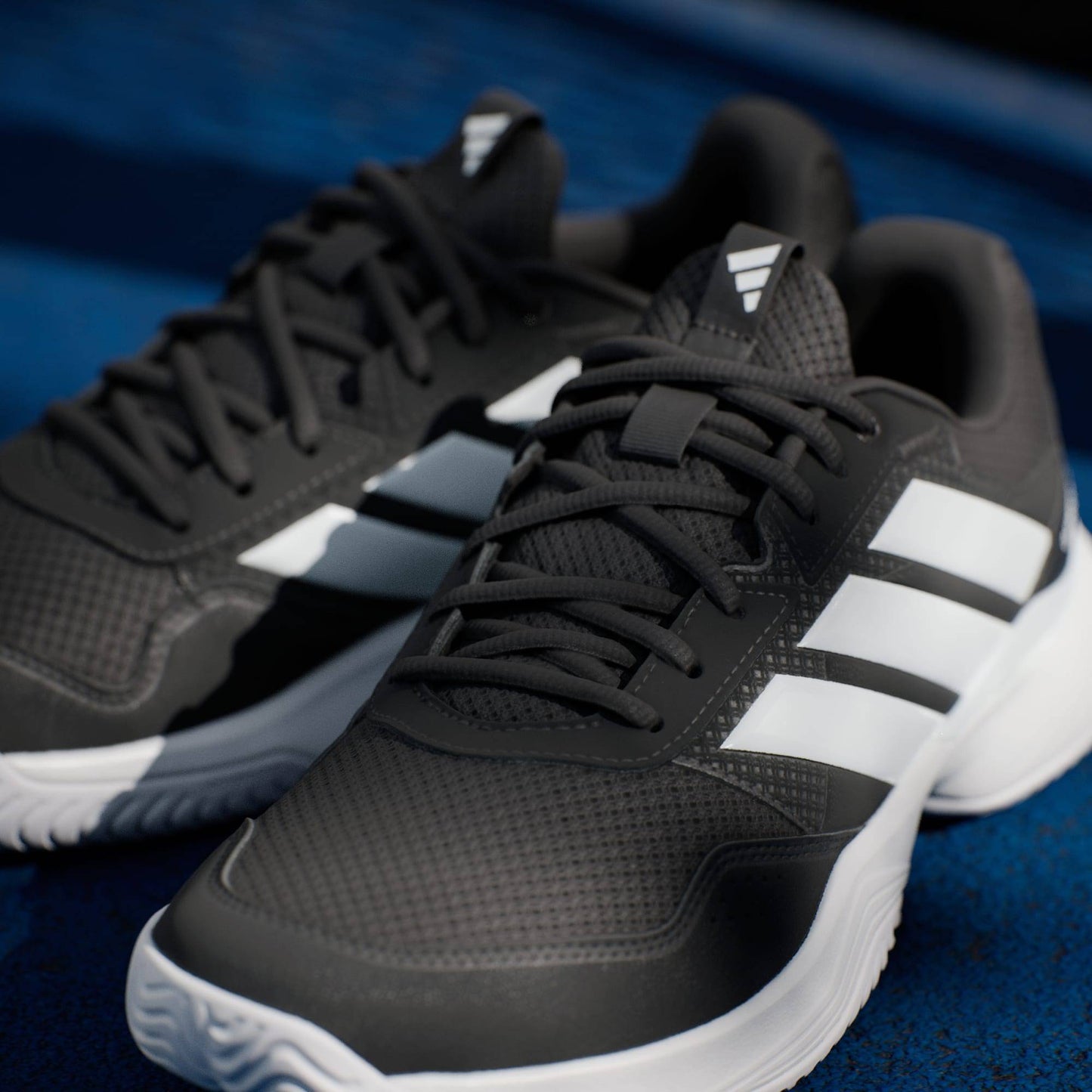 A close-up of adidas Gamecourt 3 Mens Tennis Shoes - Core Black shows black athletic sneakers with white soles and three white stripes, placed on a blue surface.