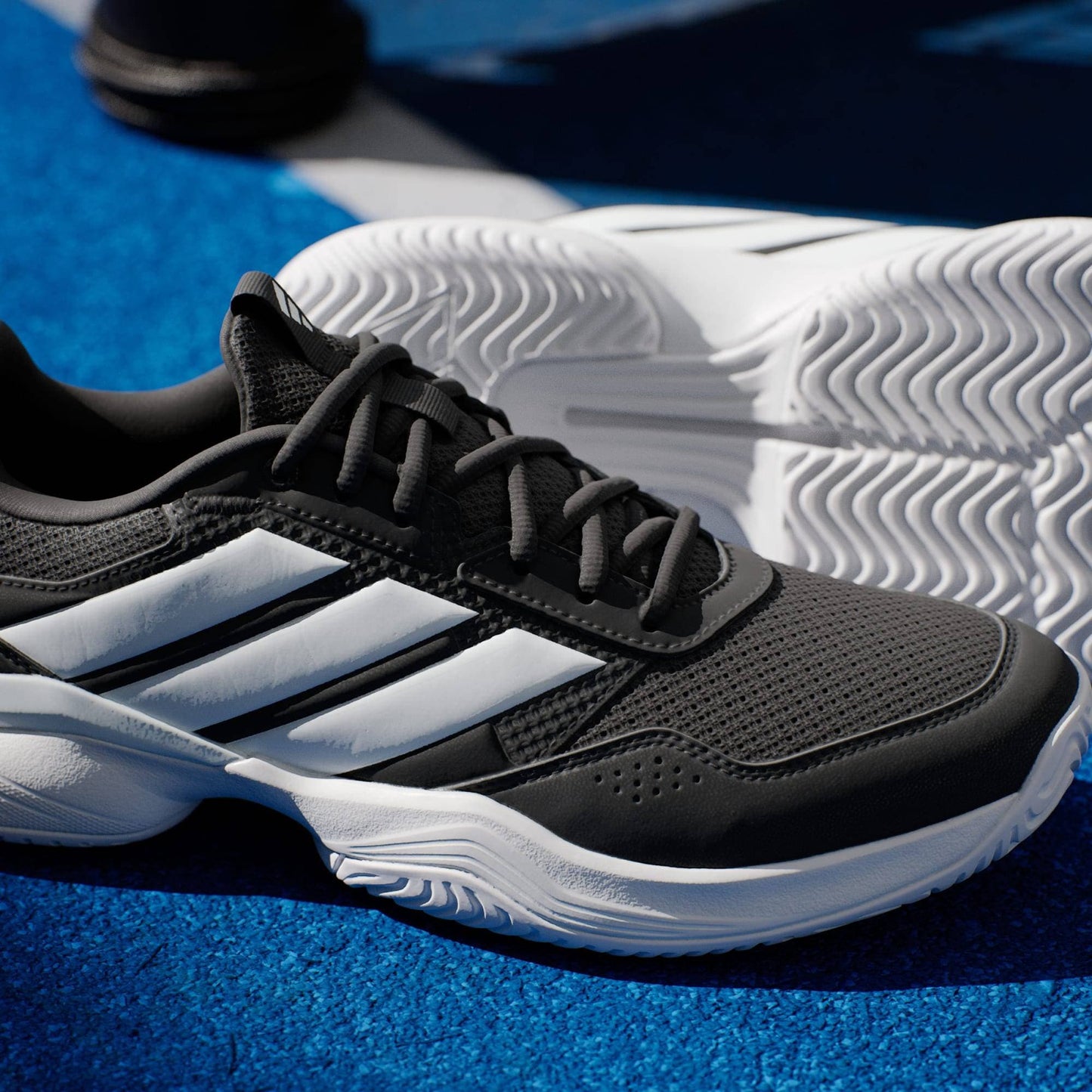 A close-up of the adidas Gamecourt 3 Mens Tennis Shoes – Core Black, featuring signature white stripes and a sturdy white sole on a vibrant blue court, with another stylish adidas tennis shoe visible in the background.