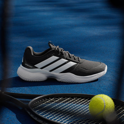 A pair of adidas Gamecourt 3 Men's Tennis Shoes in Core Black rests on a blue court next to a black tennis racket and yellow ball, with partial shadows across the scene.