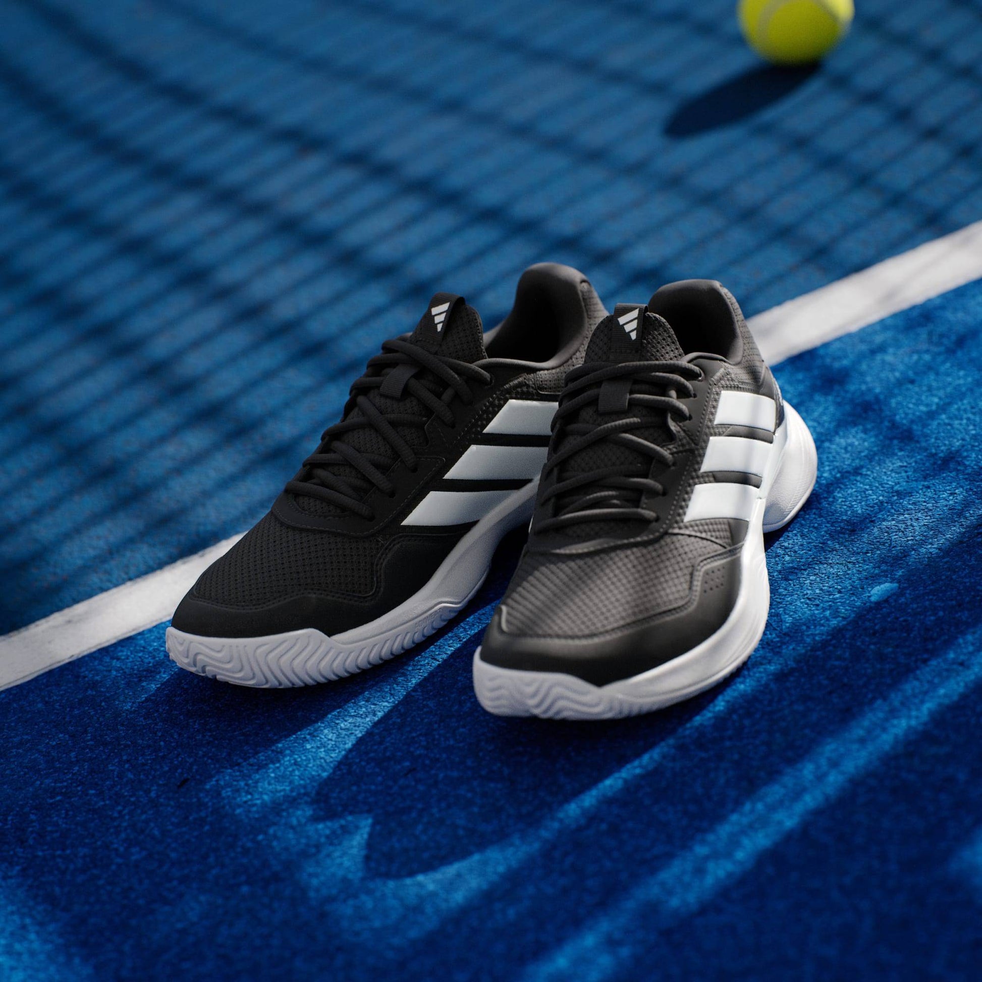 A pair of adidas Gamecourt 3 Men's Tennis Shoes in Core Black sit on a blue tennis court, with a tennis ball and the net’s shadow in the background.
