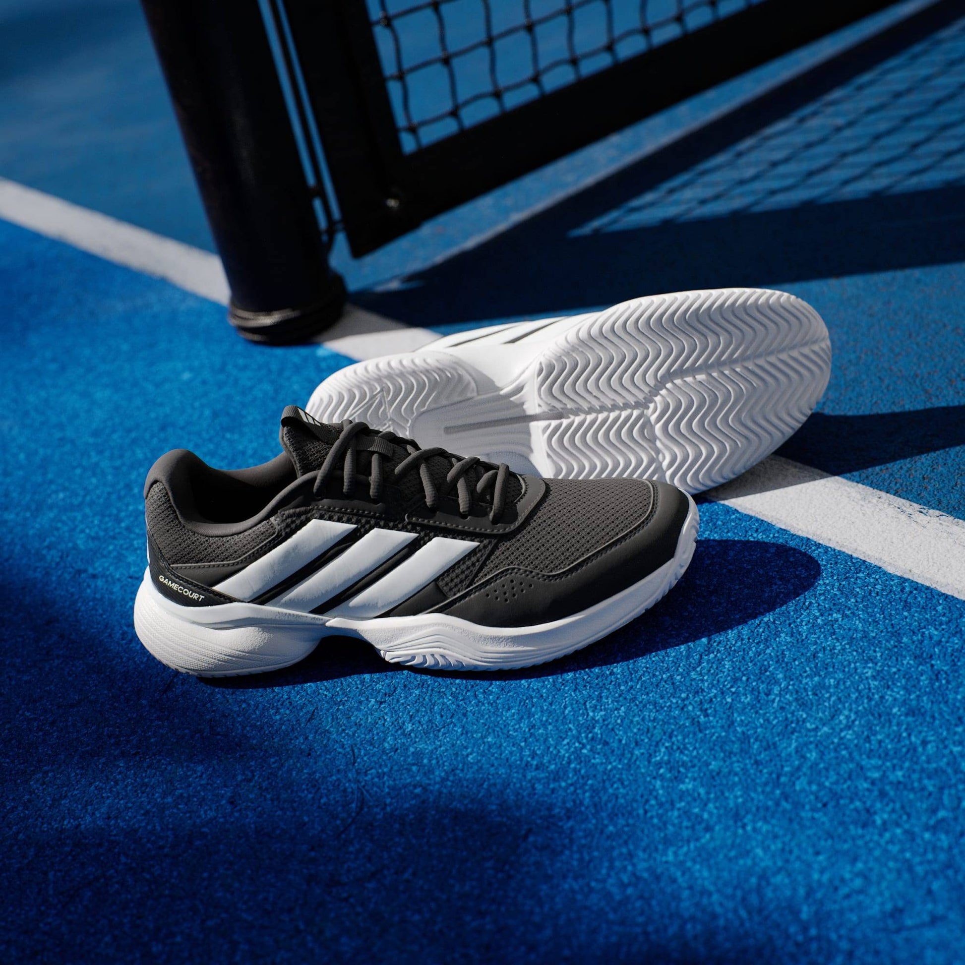 A pair of adidas Gamecourt 3 Men's Tennis Shoes in Core Black with white stripes and soles sit on a bright blue court near the net, highlighting their stylish look and lightweight design from adidas.