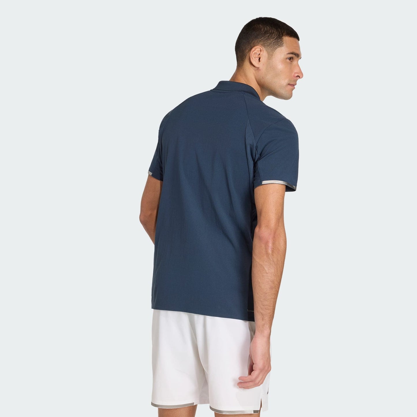 A man wearing an adidas Climacool Men's Tennis Polo Shirt in Aurora Ink with white shorts stands facing away, looking over his right shoulder against a plain white background.