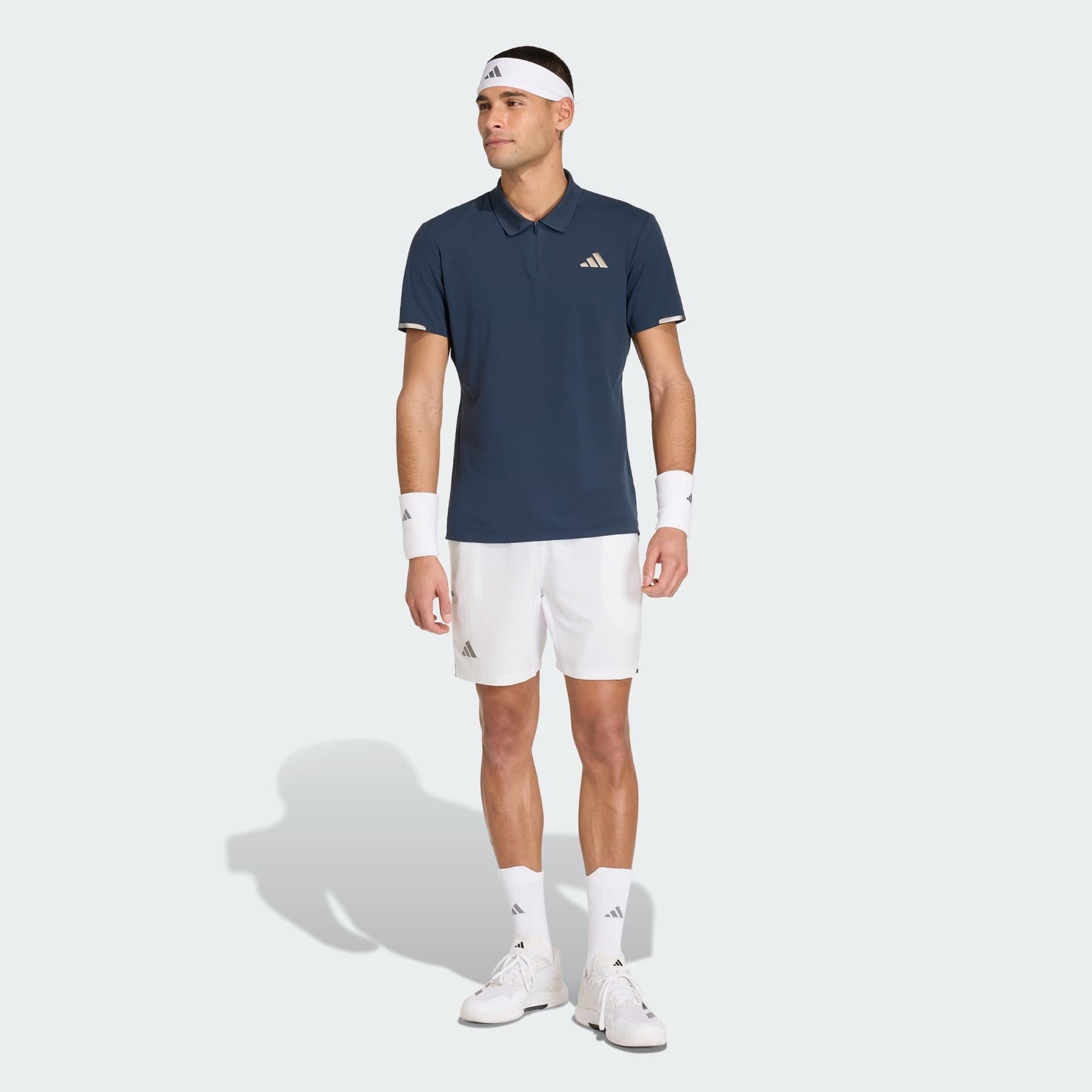 A man wearing an adidas Climacool Men's Tennis Polo Shirt in Aurora Ink, white shorts, white sneakers, socks, wristbands, and a headband stands against a plain light background.
