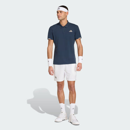 A man wearing an adidas Climacool Men's Tennis Polo Shirt in Aurora Ink, white shorts, white sneakers, socks, wristbands, and a headband stands against a plain light background.