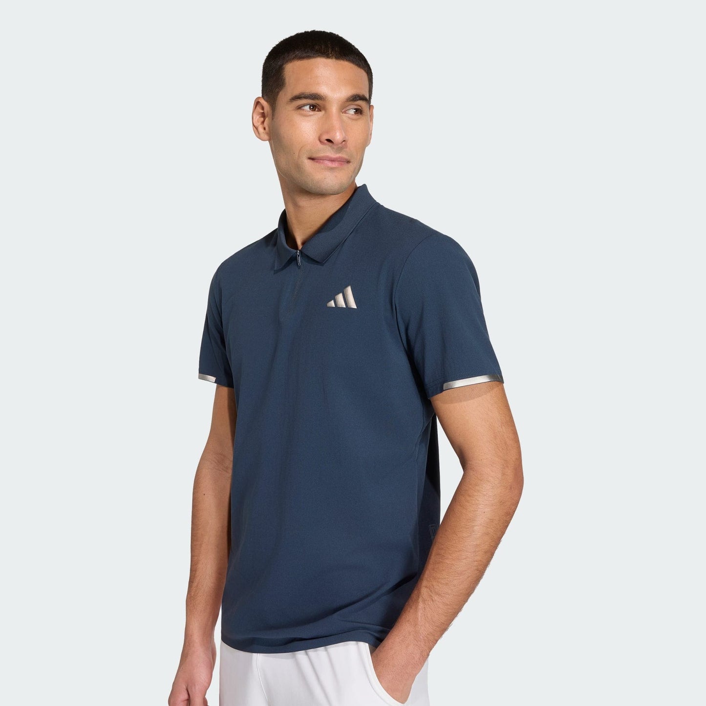 A man in an adidas Climacool Men's Tennis Polo Shirt in Aurora Ink, featuring a small white chest logo and FreeLift design, pairs it with white shorts while standing against a light background, glancing to the side.