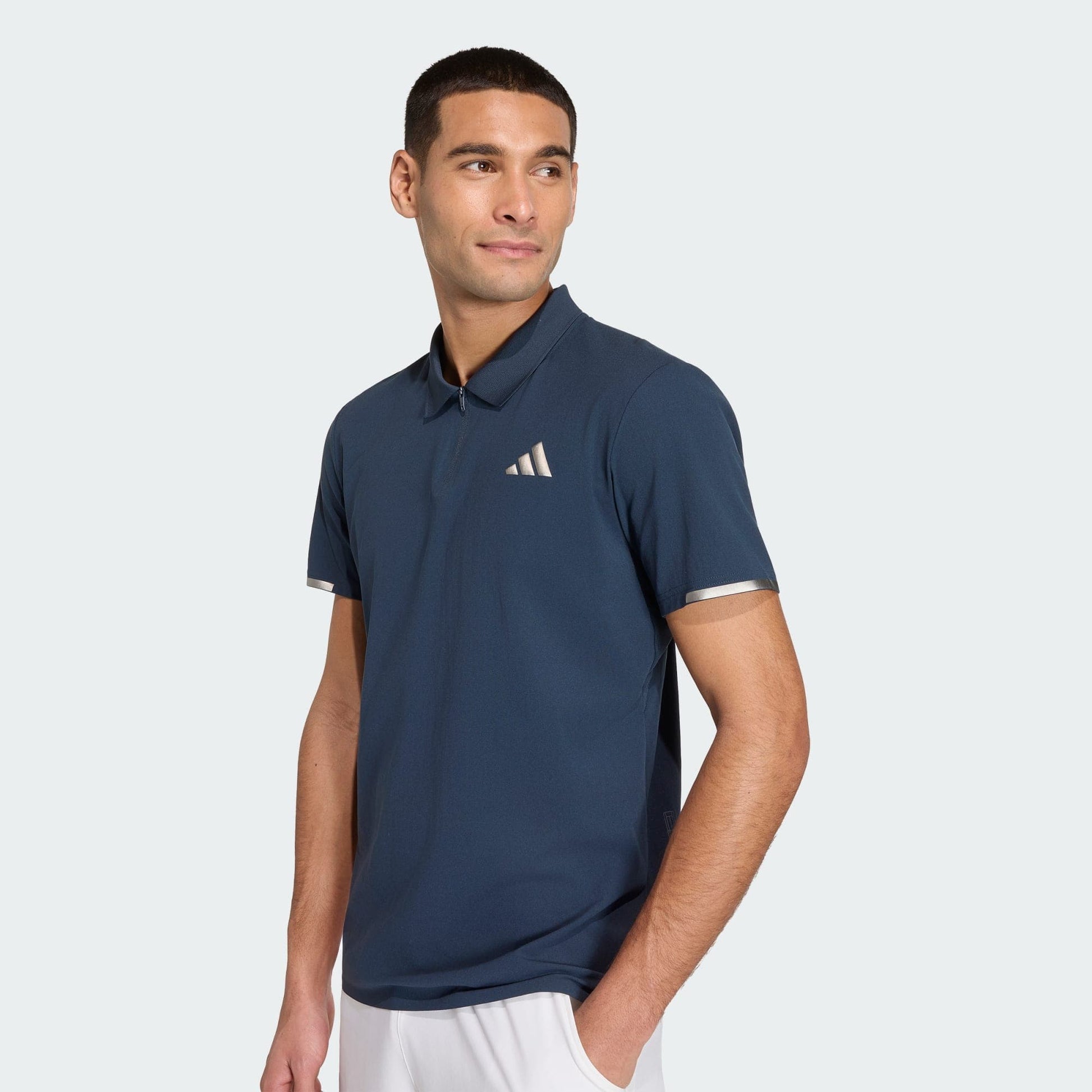 A man in an adidas Climacool Men's Tennis Polo Shirt in Aurora Ink, featuring a small white chest logo and FreeLift design, pairs it with white shorts while standing against a light background, glancing to the side.