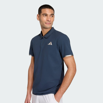 A man in an adidas Climacool Men's Tennis Polo Shirt in Aurora Ink, featuring a small white chest logo and FreeLift design, pairs it with white shorts while standing against a light background, glancing to the side.