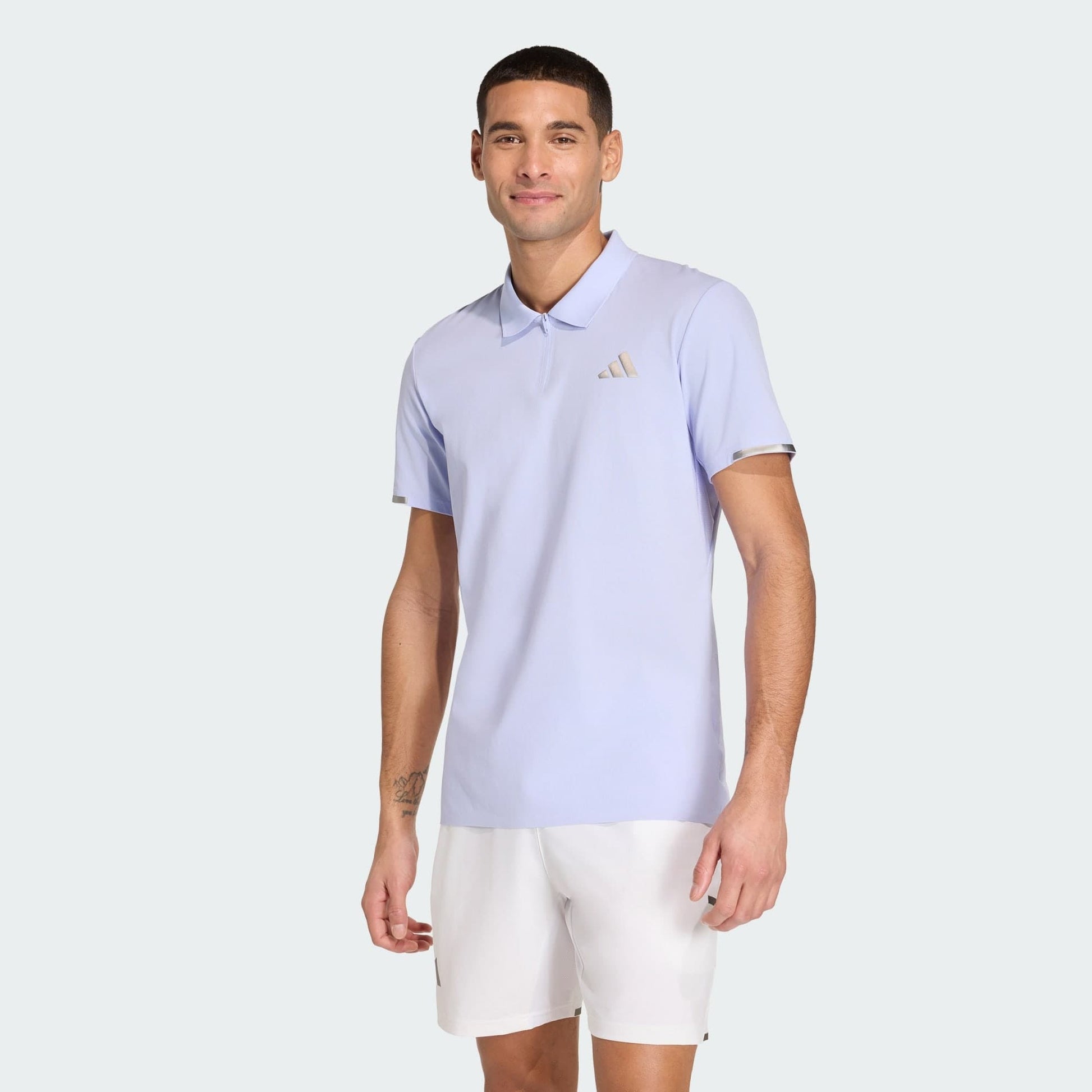 A man wearing an adidas Climacool Mens Tennis Polo Shirt in Violet Tone and white shorts stands against a plain light background, smiling at the camera.