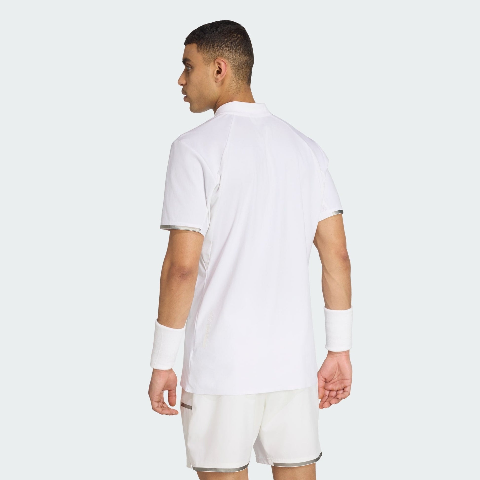 A man stands facing away, wearing an adidas Climacool Men's Tennis Polo Shirt in white, with matching shorts and wristbands, against a plain light background.