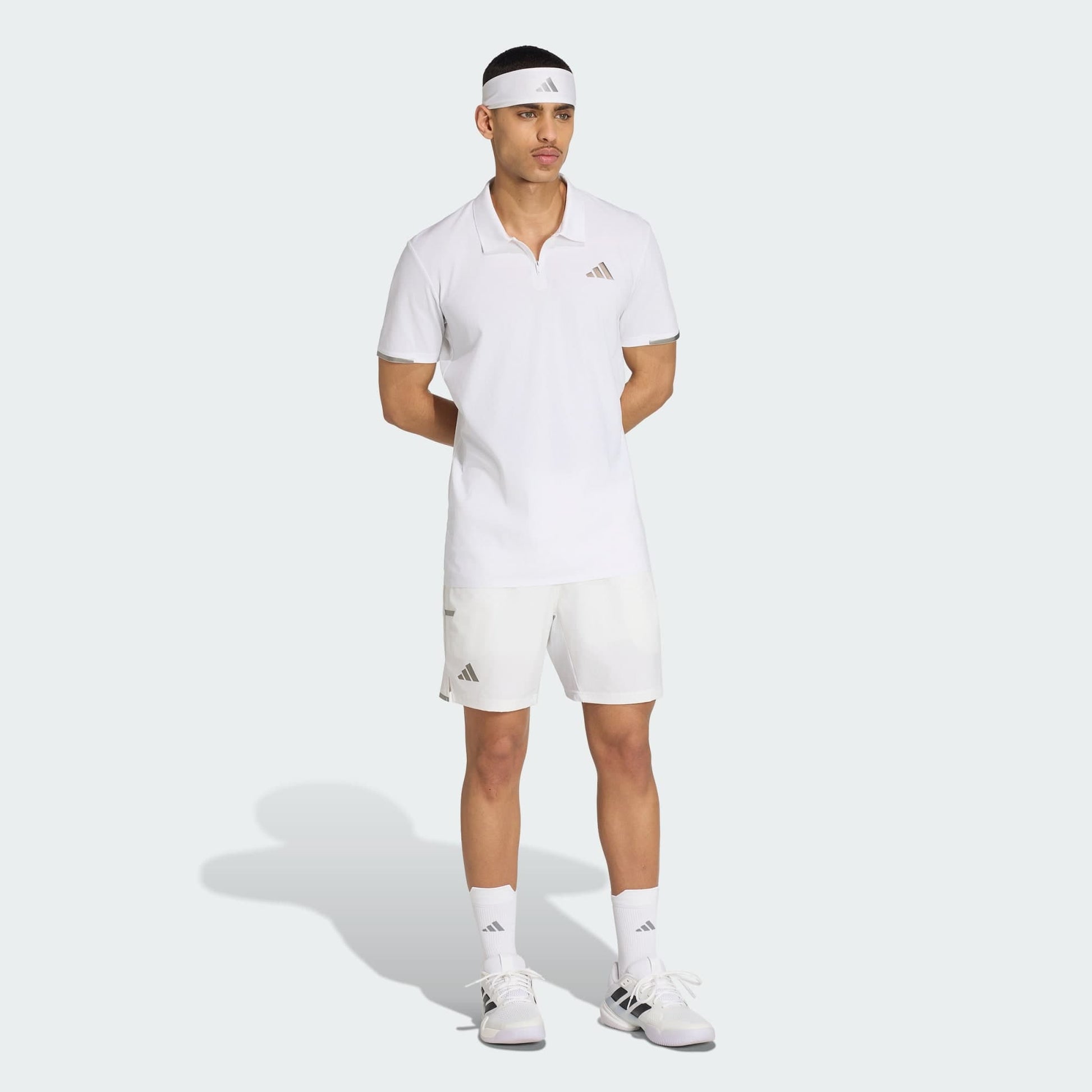A man wearing a white adidas Climacool Mens Tennis Polo Shirt, matching shorts, socks, shoes, and a headband stands with his hands behind his back against a light background.