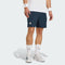 A person is shown from the waist down wearing navy adidas Climacool Men's Tennis Shorts in Aurora Ink, a white T-shirt, white adidas socks, white sneakers, and white wristbands.