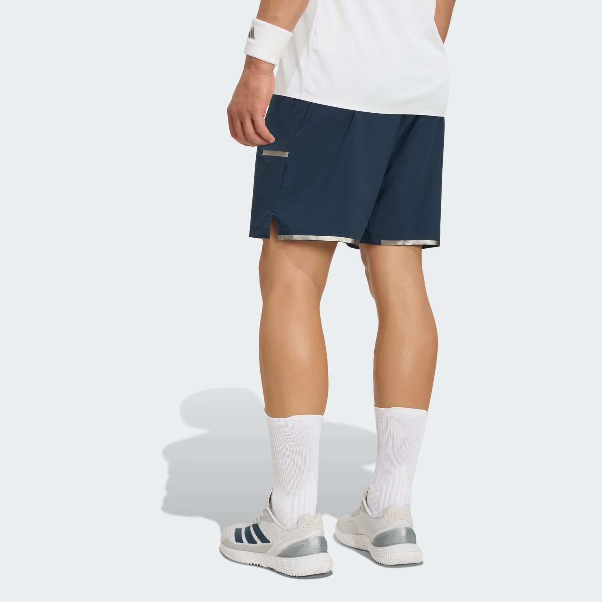A person stands facing away, wearing a white shirt, adidas Climacool Men's Tennis Shorts in Aurora Ink, white crew socks, white sneakers, and a white wristband against a plain light background.