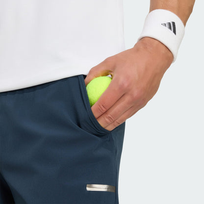 A person in adidas Climacool Men's Tennis Shorts - Aurora Ink and a white shirt slips a yellow tennis ball into their pocket, sporting a white wristband with a black logo—ready for action in these moisture-wicking tennis shorts with handy ball pockets.