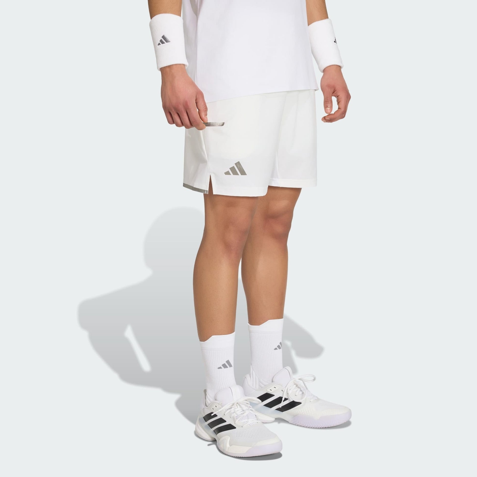 A person wearing adidas Climacool Mens Tennis Shorts in white, paired with white socks, wristbands, and black-accented white adidas sneakers, stands against a plain light background.