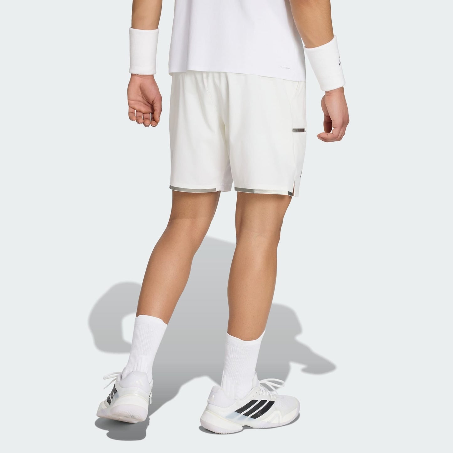 A person faces away, wearing a white shirt, adidas Climacool Men's Tennis Shorts in white by adidas, white socks, white shoes with black stripes, and white wristbands against a plain light gray background.