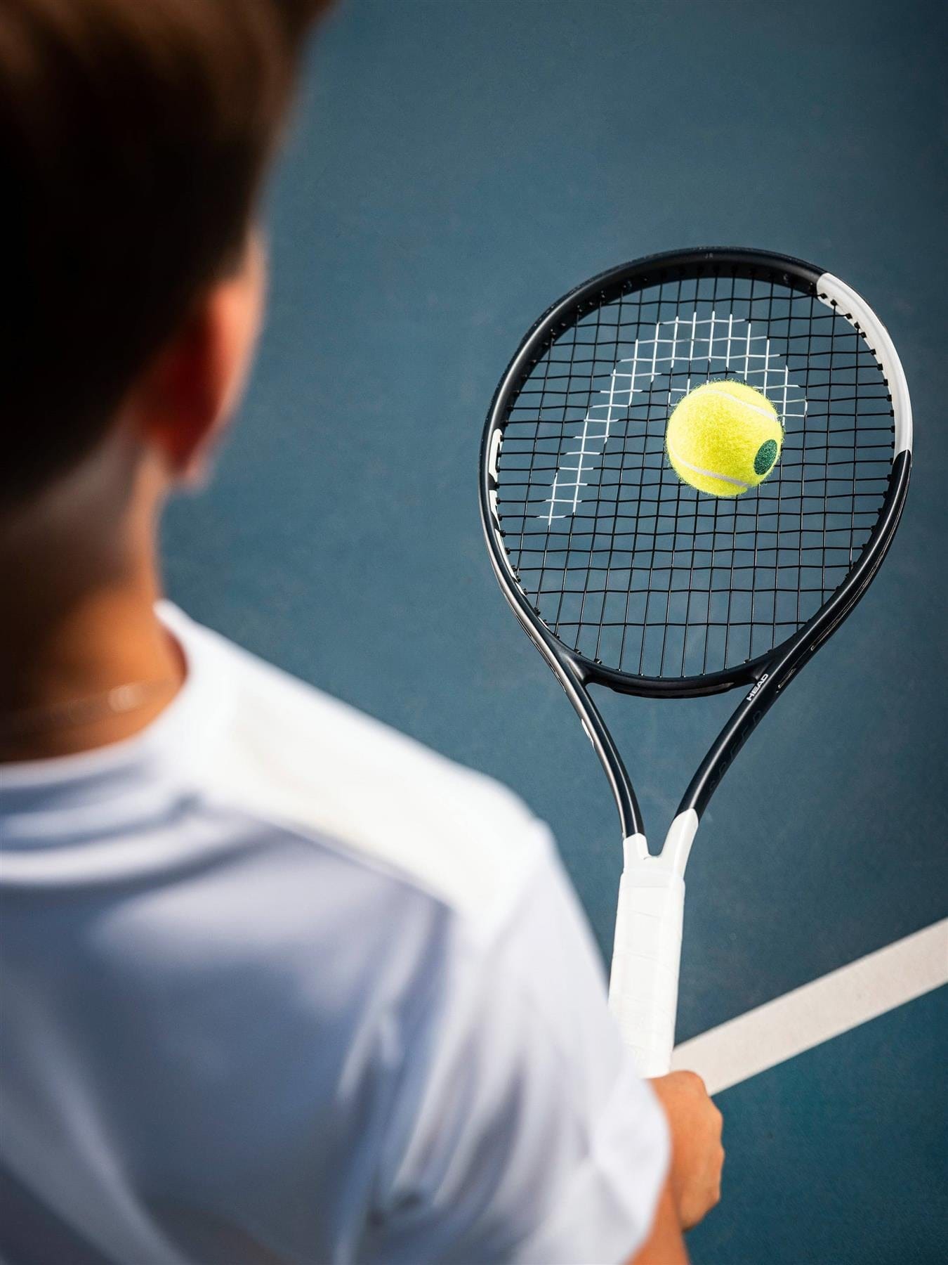 A person in a white shirt holds a HEAD Speed Junior 26 2026 Tennis Racket by HEAD, with a yellow tennis ball hovering above the junior racket on a blue court, viewed from slightly behind.
