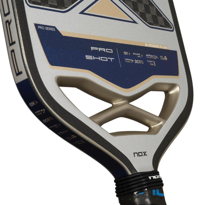 Close-up of the NOX Pro Shot Pickleball Paddle by NOX, showcasing its blue, gold, and silver design. The carbon fibre paddle features branding, spin technology details, geometric cutouts near the handle, and a black grip tape.