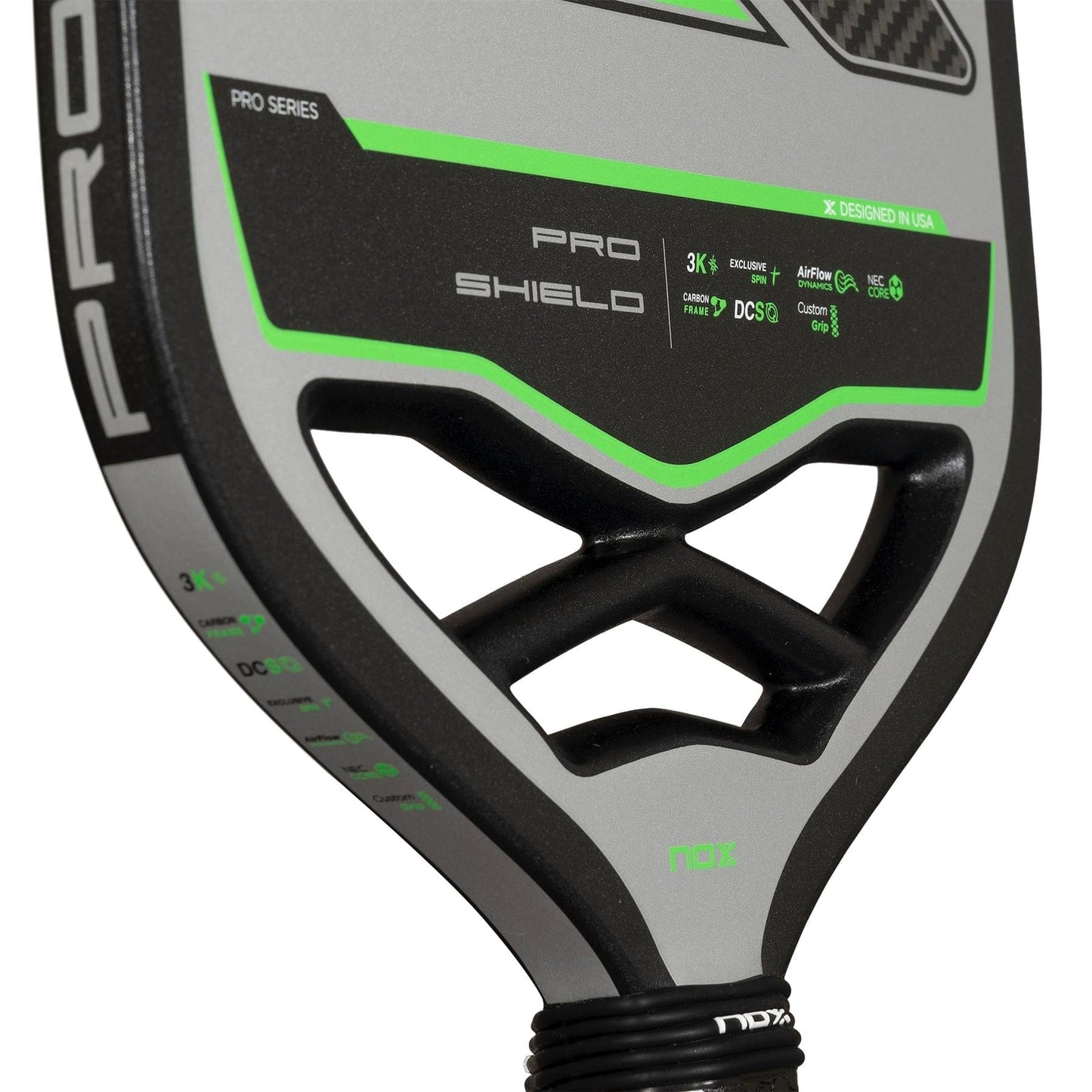 Close-up of the NOX Pro Shield Pickleball Paddle in black and gray with green accents, showing the handle and face. The paddle features "PRO SHIELD," specs, and advanced spin tech printed on its carbon fiber surface. Brand: NOX.