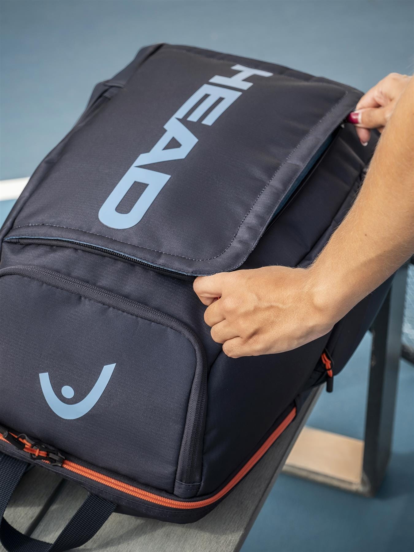 A person opens a HEAD Pro 28L Tennis Backpack - Navy, featuring a blue logo and orange zipper accents on a wooden bench, revealing its spacious racket compartment.